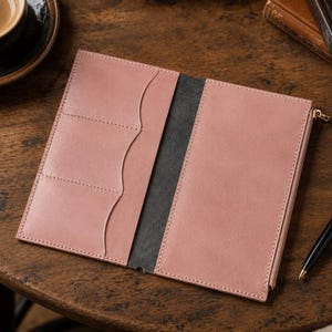 May include: A dusty rose leather wallet featuring a vertical orientation with three card slots on the left side, a central dark gray fabric spine, and a zippered compartment on the right side, finished with a small gold-toned metal zipper pull.