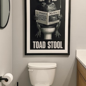 Puede incluir: Impresión en blanco y negro de una rana con sombrero de copa leyendo un periódico titulado "Toadly News". La impresión está encima de un inodoro blanco. El texto "TOAD STOOL" está en la parte inferior de la impresión.