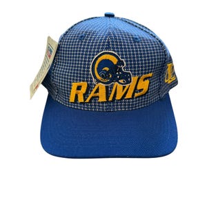 May include: A blue and white houndstooth patterned baseball cap featuring an embroidered yellow and blue Rams logo with the text RAMS in yellow. The cap has a solid blue brim and a white paper tag attached to the side panel of the crown.