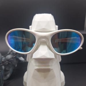 May include: White Oakley sunglasses with blue reflective lenses rest on a white geometric sculpture. The sculpture has a stylized face. The sunglasses have the Oakley logo on the frame.