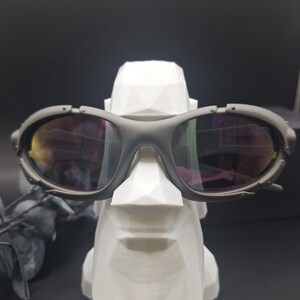 May include: Gray sunglasses with reflective lenses rest on a white, geometric sculpture of a face. The sunglasses have a thick frame and a sporty design. The sculpture is a stylized representation of a face.