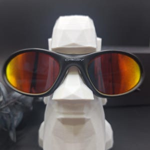 May include: Black Oakley sunglasses with orange and yellow lenses rest on a white geometric sculpture. The sunglasses have the Oakley logo in white on the frame. The sculpture is a stylized face.