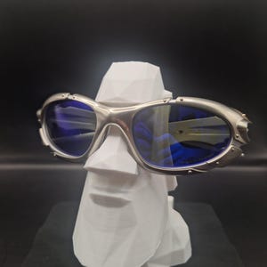 May include: Silver-framed sunglasses with blue lenses rest on a white, geometric sculpture of a face. The sunglasses have a sporty design with a metallic finish. The background is a solid black.