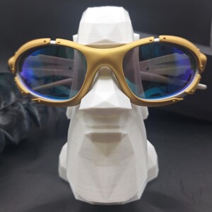May include: Gold-framed sunglasses with blue lenses rest on a white, geometric sculpture of a face. The sunglasses have a sporty design with a gold finish and blue-tinted lenses. The sculpture is white and has a modern, angular design.