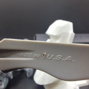May include: Close-up of a gray plastic tool with the text "MADE IN U.S.A." printed on it. The tool has a small emblem next to the text. A white, geometric shape is in the background.