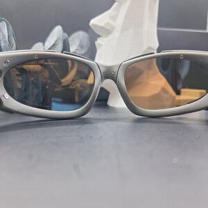 May include: Grey sunglasses with oval lenses and a matte finish. The sunglasses have a sporty design with visible hinges and a subtle brow bar. The lenses have a gradient tint, transitioning from a darker shade at the top to a lighter hue at the bottom.