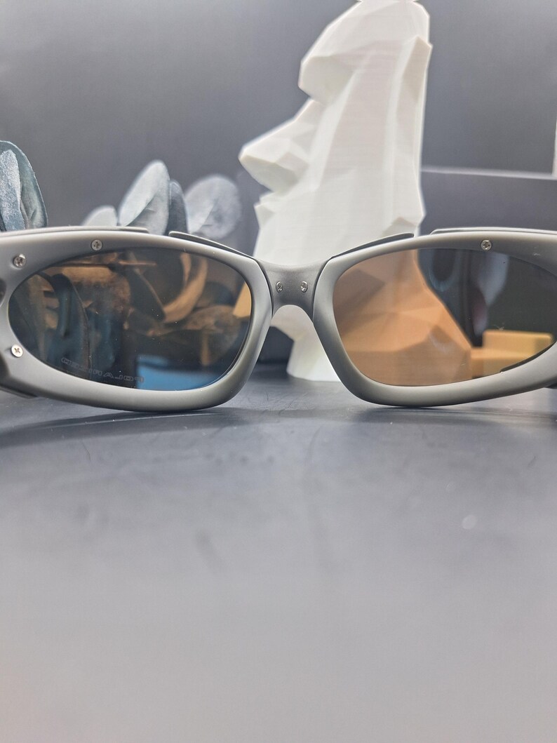 May include: Grey sunglasses with oval lenses and a matte finish. The sunglasses have a sporty design with visible hinges and a subtle brow bar. The lenses have a gradient tint, transitioning from a darker shade at the top to a lighter hue at the bottom.