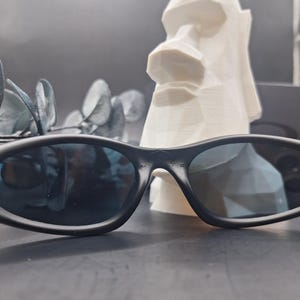 May include: Black sunglasses with dark blue lenses are in the foreground. Behind them is a white, geometric sculpture of a Moai head. The sunglasses have a sleek, modern design.