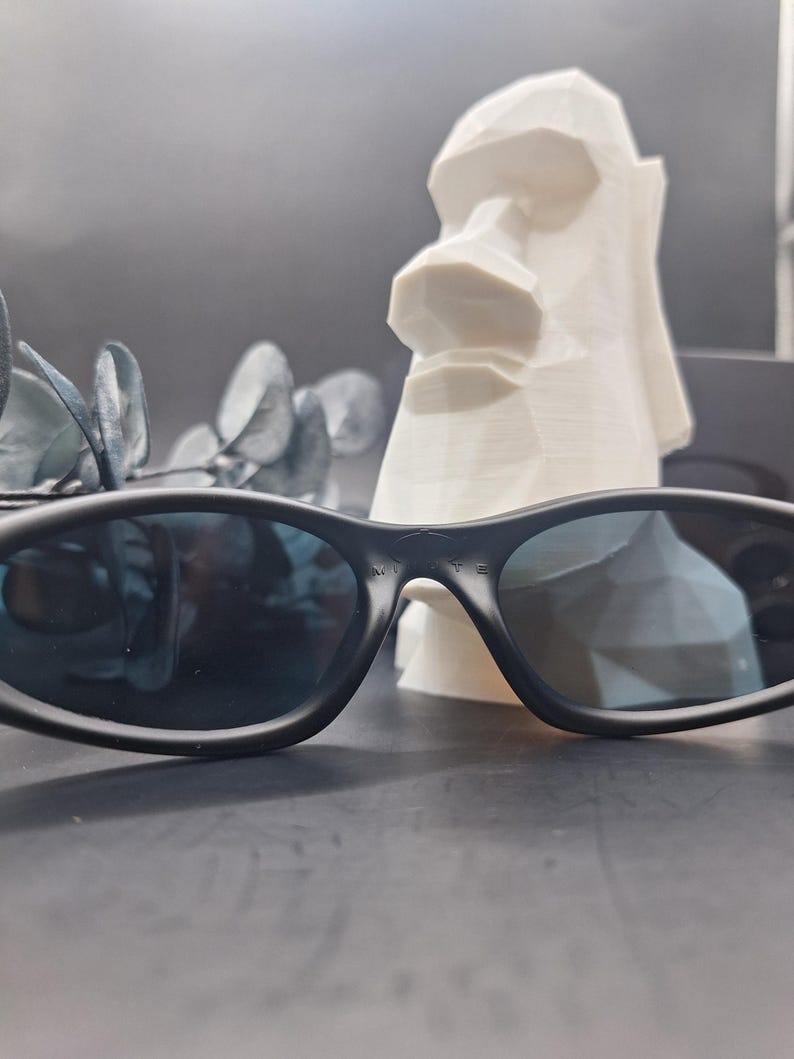 May include: Black sunglasses with dark blue lenses are in the foreground. Behind them is a white, geometric sculpture of a Moai head. The sunglasses have a sleek, modern design.