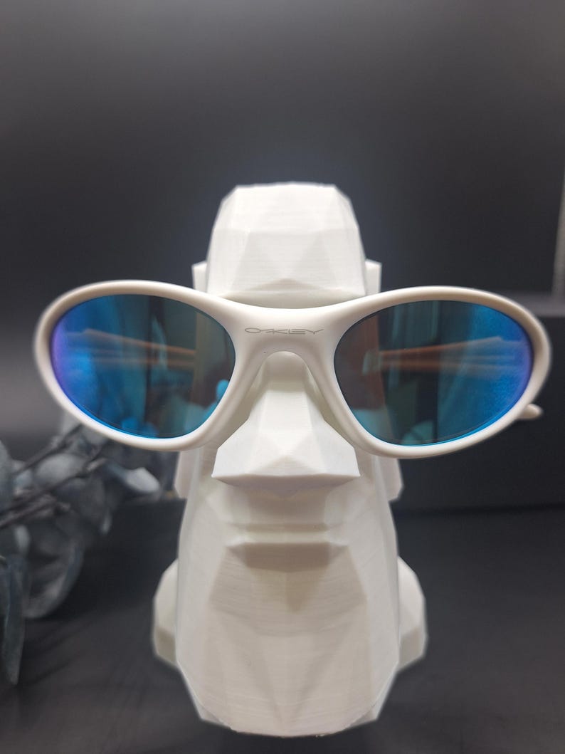 May include: White Oakley sunglasses with blue reflective lenses rest on a white geometric sculpture. The sculpture has a stylized face. The sunglasses have the Oakley logo on the frame.