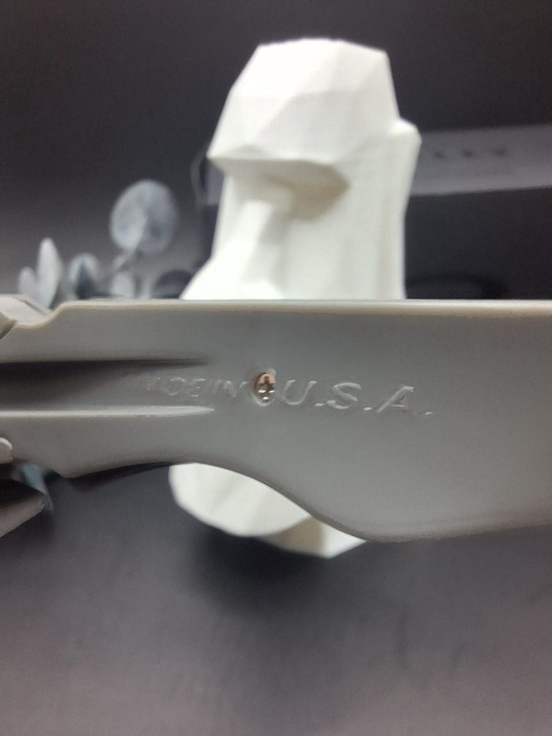 Może przedstawiać: Szare plastikowe narzędzie z napisem "MADE IN U.S.A." znajduje się na pierwszym planie. Biała, geometryczna rzeźba głowy znajduje się w tle. Rzeźba ma płaską g&oacute;rę i kanciaste cechy.