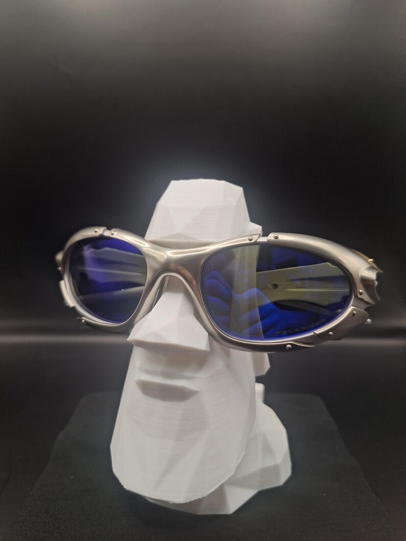 May include: Silver-framed sunglasses with blue lenses rest on a white, geometric sculpture of a face. The sunglasses have a sporty design with a metallic finish. The background is a solid black.