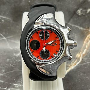 Könnte beinhalten: Eine Oakley-Uhr aus Edelstahl mit einem schwarzen Kautschukarmband. Das Zifferblatt der Uhr ist rot mit schwarzen Hilfszifferblättern und weißen Markierungen. Die Uhr wird auf einem weißen Ständer präsentiert.