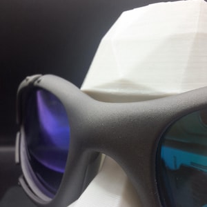 Op de afbeelding: Close-up van een zwarte sportbril met blauw getinte glazen. De bril rust op een witte, geometrisch gevormde standaard. De glazen reflecteren een verloop van blauwe en paarse tinten en het montuur heeft een matte afwerking.