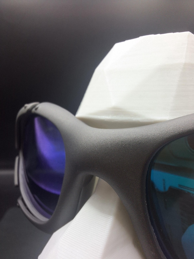 Op de afbeelding: Close-up van een zwarte sportbril met blauw getinte glazen. De bril rust op een witte, geometrisch gevormde standaard. De glazen reflecteren een verloop van blauwe en paarse tinten en het montuur heeft een matte afwerking.