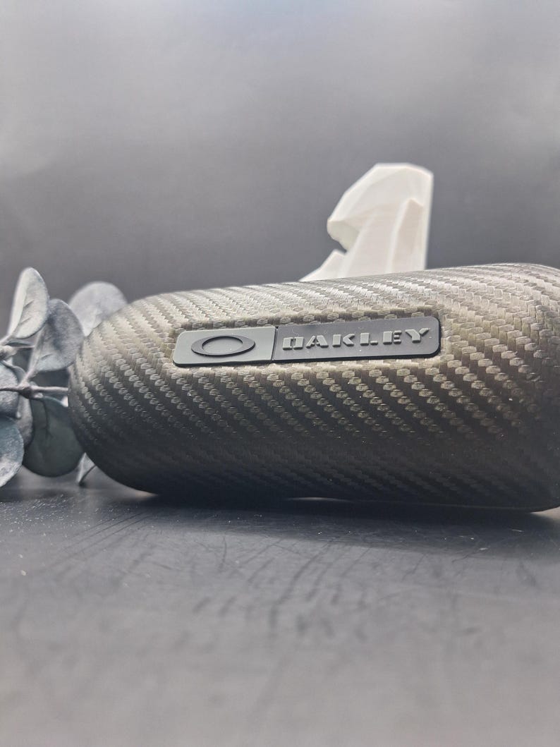 Może przedstawiać: Owalne etui na okulary z fakturą wł&oacute;kna węglowego i wytłoczonym logo marki "OAKLEY". Etui jest w kolorze ciemnoszarym. Gałązka eukaliptusa i biała chusteczka znajdują się w tle.