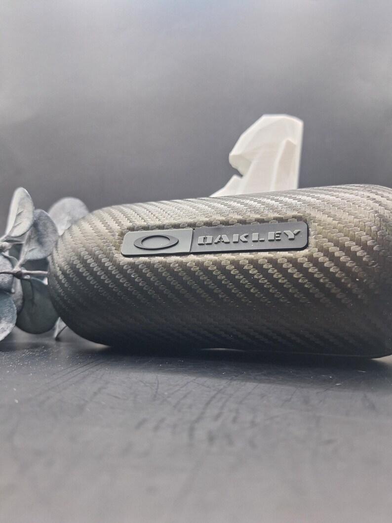 May include: A textured, carbon fiber-style Oakley sunglasses case. The case is oval-shaped with the Oakley logo and brand name in a dark gray color. The case is on a dark surface with a sprig of greenery.