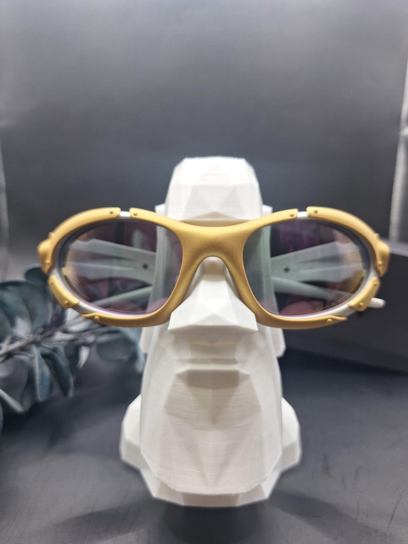 May include: Gold-framed sunglasses with oval lenses rest on a white geometric sculpture. The sunglasses have a gold bridge and temples, and the lenses have a slight tint. The sculpture is a stylized head.