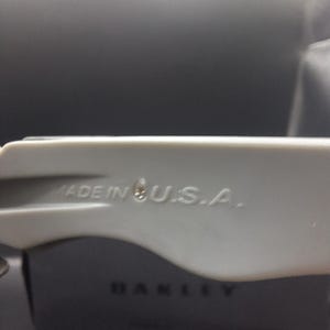 Op de afbeelding: Close-up van een lichtgrijze Oakley zonnebrilarm met de tekst "MADE IN U.S.A.". Het Oakley-logo is zichtbaar op de zwarte case op de achtergrond. De zonnebrilarm is gemaakt van een glad, mat materiaal.