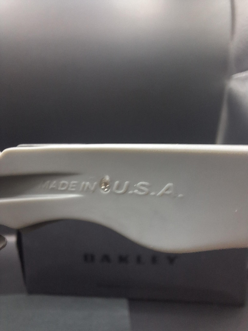 Op de afbeelding: Close-up van een lichtgrijze Oakley zonnebrilarm met de tekst "MADE IN U.S.A.". Het Oakley-logo is zichtbaar op de zwarte case op de achtergrond. De zonnebrilarm is gemaakt van een glad, mat materiaal.