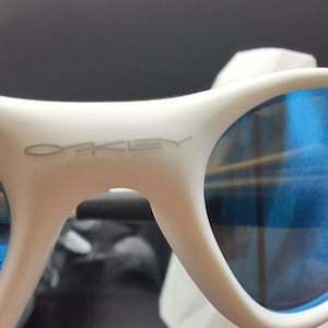 May include: White sunglasses with blue lenses. The brand name "Oakley" is visible on the frame. The sunglasses have a modern design and are suitable for outdoor activities.