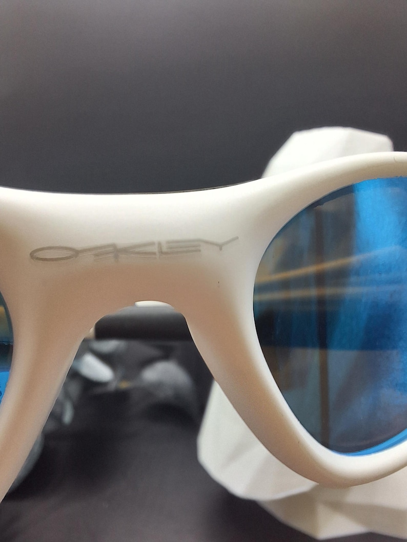 May include: White sunglasses with blue lenses. The brand name "Oakley" is visible on the frame. The sunglasses have a modern design and are suitable for outdoor activities.