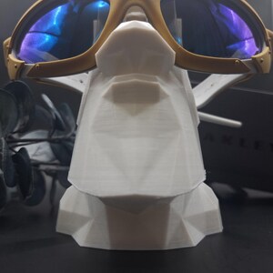 May include: Gold-framed sunglasses with blue reflective lenses rest on a white geometric display stand. The sunglasses have a sporty design with a double bridge and visible hinges. The stand is a modern, angular design.