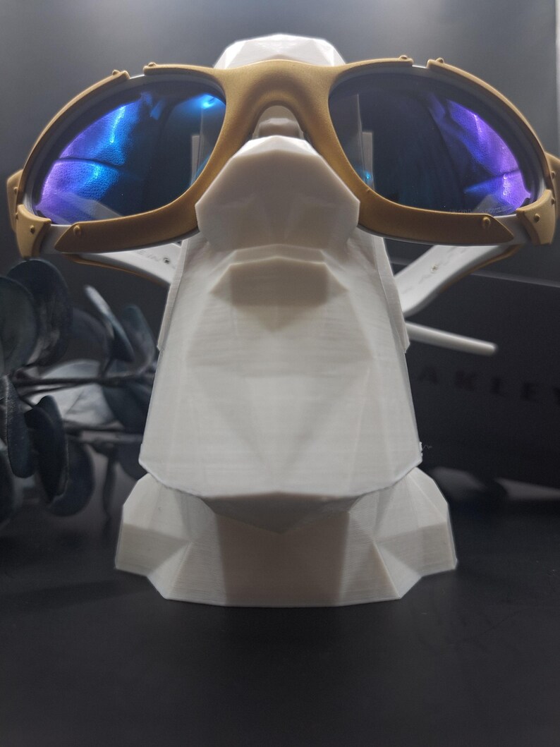 May include: Gold-framed sunglasses with blue reflective lenses rest on a white geometric display stand. The sunglasses have a sporty design with a double bridge and visible hinges. The stand is a modern, angular design.