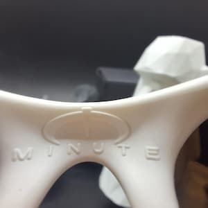 May include: Close-up of a white plastic object with the word "MINUTE" embossed on it. The object has a curved design with a circular emblem above the text. The background is dark and out of focus.