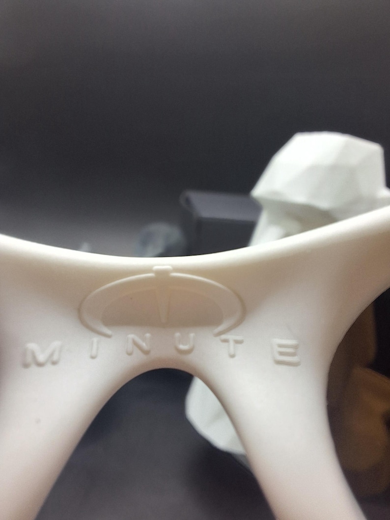 May include: Close-up of a white plastic object with the word "MINUTE" embossed on it. The object has a curved design with a circular emblem above the text. The background is dark and out of focus.