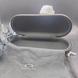 May include: An open, oval glasses case with a textured, dark grey exterior. Inside, the case is lined with black fabric. A black drawstring pouch with the "Oakley" logo is visible, along with a white tissue.