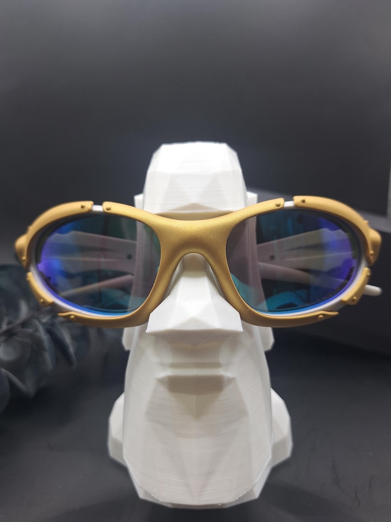 May include: Gold-framed sunglasses with blue lenses rest on a white, geometric sculpture of a face. The sunglasses have a sporty design with a gold finish and blue-tinted lenses. The sculpture is white and has a modern, angular design.