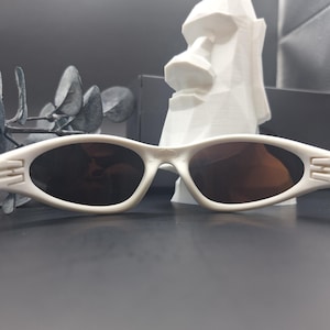 May include: White oval sunglasses with dark lenses. The sunglasses have a sleek, modern design with a subtle detail on the temples. The image also features a white Moai statue and decorative foliage in the background.