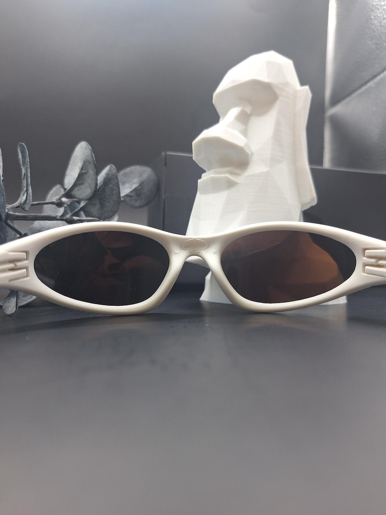 May include: White oval sunglasses with dark lenses. The sunglasses have a sleek, modern design with a subtle detail on the temples. The image also features a white Moai statue and decorative foliage in the background.