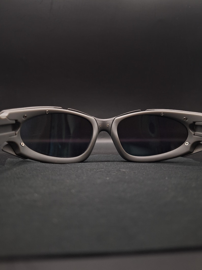 Op de afbeelding: Close-up van een grijze zonnebril met donkere glazen. De zonnebril heeft een wrap-around design met een dik montuur en kleine metalen accenten. De glazen zijn ovaal van vorm en reflecteren de omgeving. De zonnebril staat tegen een donkere achtergrond.