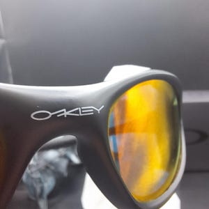 May include: Close-up of black sunglasses with the brand name "OAKLEY" in white. The lenses are a vibrant yellow-orange color, reflecting light. The sunglasses are resting on a white surface.