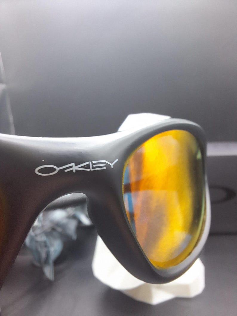 May include: Close-up of black sunglasses with the brand name "OAKLEY" in white. The lenses are a vibrant yellow-orange color, reflecting light. The sunglasses are resting on a white surface.