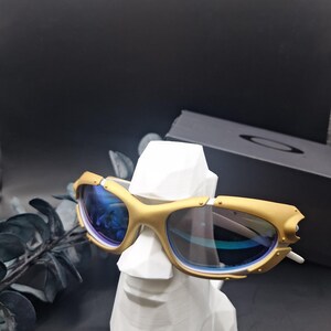 May include: Gold-framed sunglasses with blue lenses rest on a white, geometric bust. The sunglasses have a unique design with a gold finish and a sleek, modern aesthetic. The background is dark, highlighting the sunglasses.