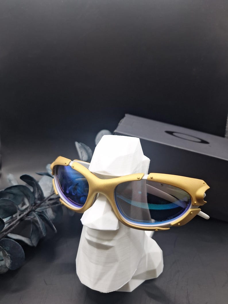 May include: Gold-framed sunglasses with blue lenses rest on a white, geometric bust. The sunglasses have a unique design with a gold finish and a sleek, modern aesthetic. The background is dark, highlighting the sunglasses.