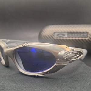 Puede incluir: Gafas de sol plateadas con lentes azules. Las gafas tienen un dise&ntilde;o angular &uacute;nico con un acabado met&aacute;lico. En el fondo se ve un estuche a juego con el nombre de la marca.