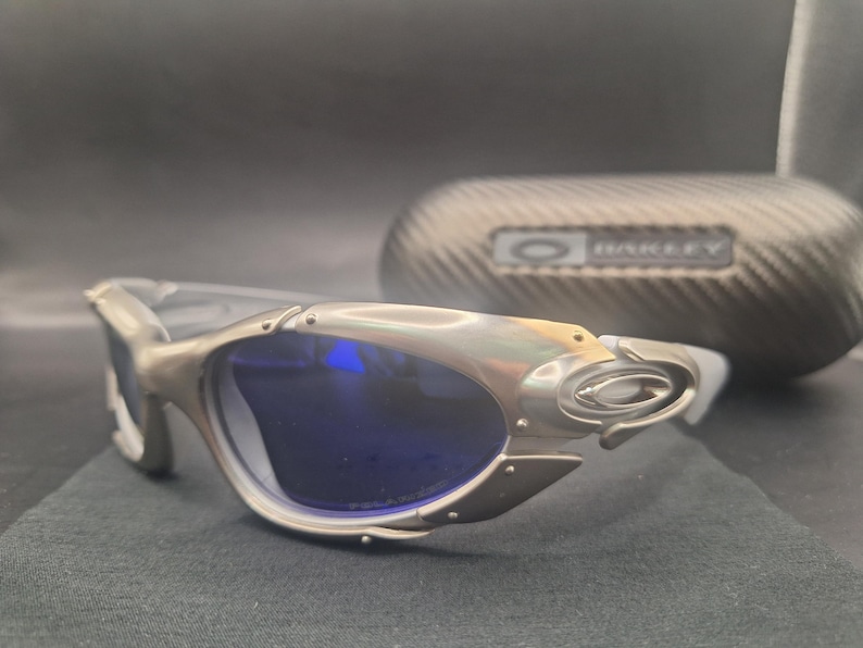 Puede incluir: Gafas de sol plateadas con lentes azules. Las gafas tienen un dise&ntilde;o angular &uacute;nico con un acabado met&aacute;lico. En el fondo se ve un estuche a juego con el nombre de la marca.