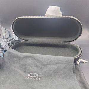 May include: An open, oval-shaped black glasses case with a textured exterior. Inside, the case is lined with black fabric. A black Oakley branded soft case is in front of the glasses case.