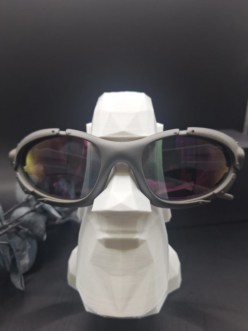May include: Gray sunglasses with reflective lenses rest on a white, geometric sculpture of a face. The sunglasses have a thick frame and a sporty design. The sculpture is a stylized representation of a face.