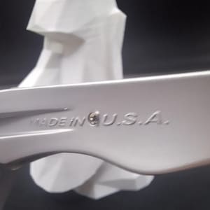 Puede incluir: Primer plano de una llave inglesa plateada con el texto "MADE IN U.S.A." grabado en el lateral. Una escultura geom&eacute;trica blanca en el fondo, sobre un fondo oscuro.