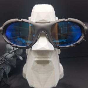 May include: Dark grey sunglasses with blue lenses are displayed on a white, geometric face sculpture. The sunglasses feature an oval frame and lenses. The sculpture is white with a faceted design, giving it a modern appearance.