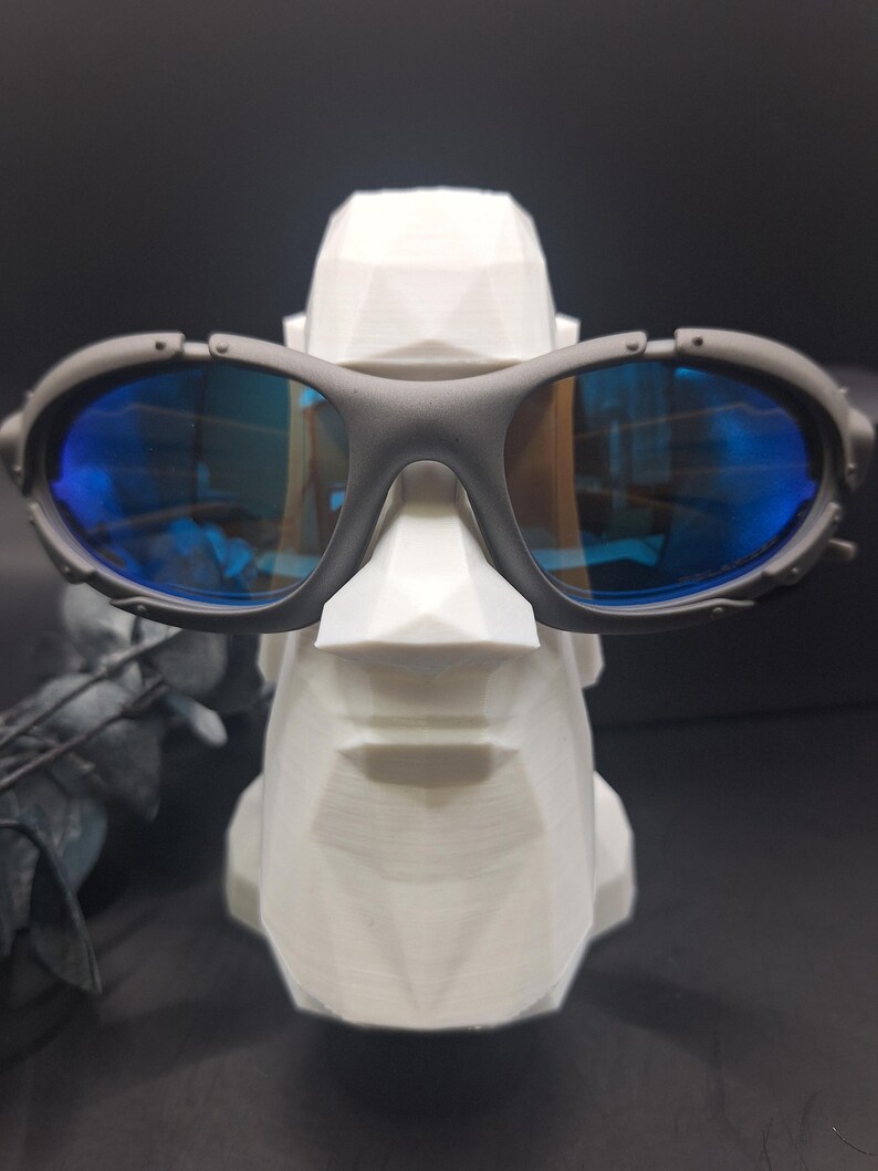 May include: Dark grey sunglasses with blue lenses are displayed on a white, geometric face sculpture. The sunglasses feature an oval frame and lenses. The sculpture is white with a faceted design, giving it a modern appearance.