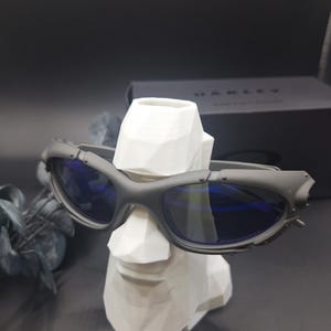 Może przedstawiać: Ciemnoszare okulary przeciwsłoneczne Oakley z niebieskimi soczewkami spoczywają na białym geometrycznym popiersiu. Okulary mają futurystyczny design z widocznymi zawiasami i matowym wykończeniem. Czarne pudełko z logo Oakley w tle.