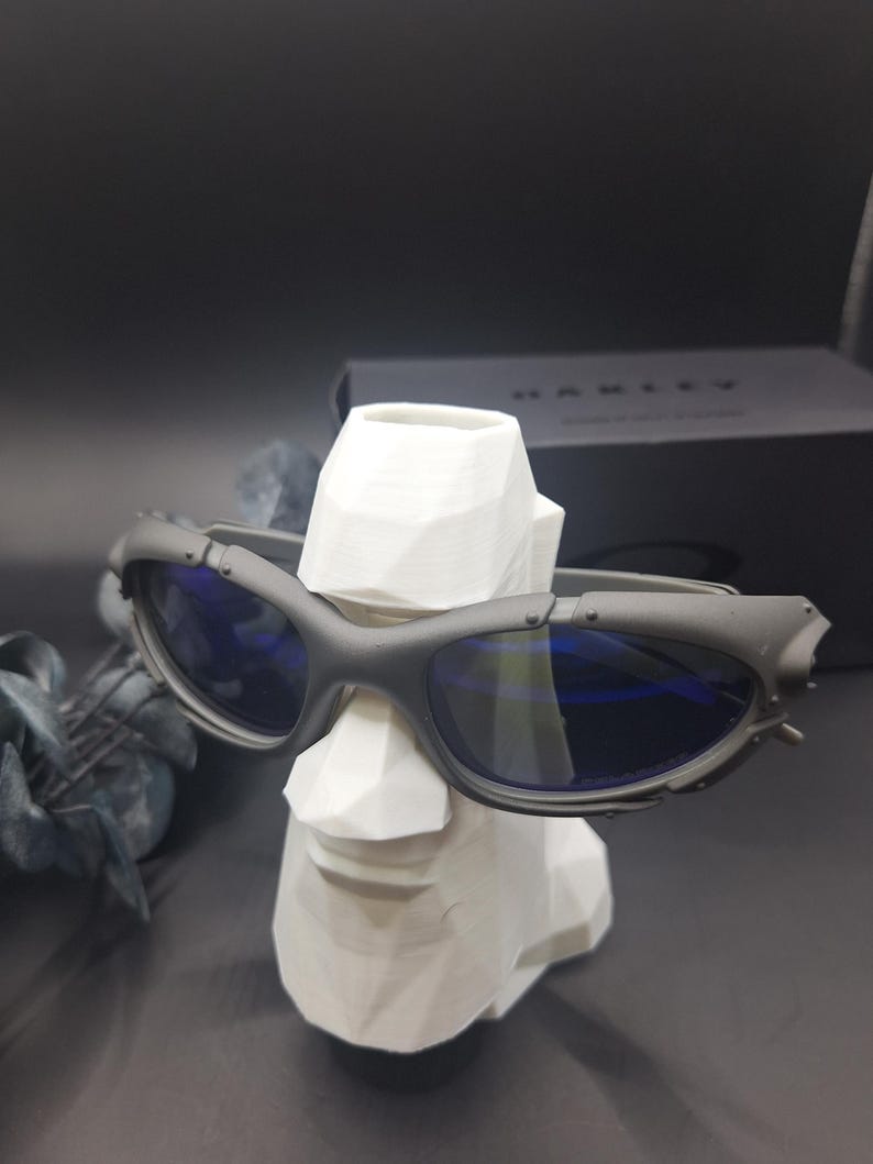Może przedstawiać: Ciemnoszare okulary przeciwsłoneczne Oakley z niebieskimi soczewkami spoczywają na białym geometrycznym popiersiu. Okulary mają futurystyczny design z widocznymi zawiasami i matowym wykończeniem. Czarne pudełko z logo Oakley w tle.