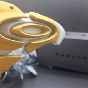 May include: Close-up of a white and gold sports eyewear frame. The frame features a gold-colored oval design with a white base. The word "OAKLEY" is visible on the side of a dark gray box in the background.