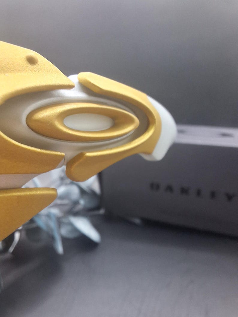 May include: Close-up of a white and gold sports eyewear frame. The frame features a gold-colored oval design with a white base. The word "OAKLEY" is visible on the side of a dark gray box in the background.